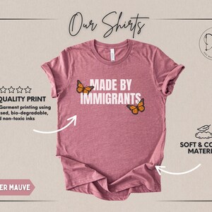 Pu&ograve; includere: Una t-shirt color malva m&eacute;lange con la scritta bianca MADE BY IMMIGRANTS centrata sul petto. Due farfalle monarca arancioni e nere sono posizionate vicino al testo, una a sinistra della parola MADE e una a destra di IMMIGRANTS.