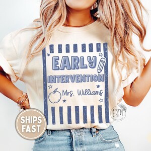 May include: An ivory t-shirt featuring a blue graphic design with vertical stripes, the text "EARLY INTERVENTION", a pencil icon, an apple outline, small stars, and the name "Mrs. Williams" printed in a script font on the front center.