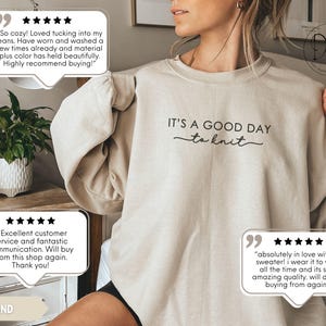 May include: A sand-colored sweatshirt with the text "IT'S A GOOD DAY to knit" in black script. The image includes customer reviews with five-star ratings. The sweatshirt is a casual, comfortable garment.
