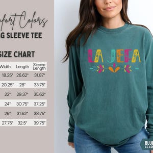 May include: Teal long-sleeve tee with the word "LA JEFA" in colourful letters. The shirt has a size chart with measurements in inches. The text "Comfort Colors" and "BLUE SPRUCE CC LONG SLEEVE TEE" are also visible.