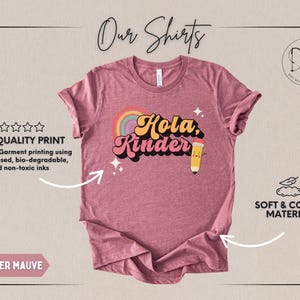 May include: A heather mauve short-sleeve t-shirt featuring a graphic print with the text "Hola, Kinder" in a retro font. The design includes a colorful rainbow, two small four-pointed stars, and a cartoon pencil with a smiling face on the right side.