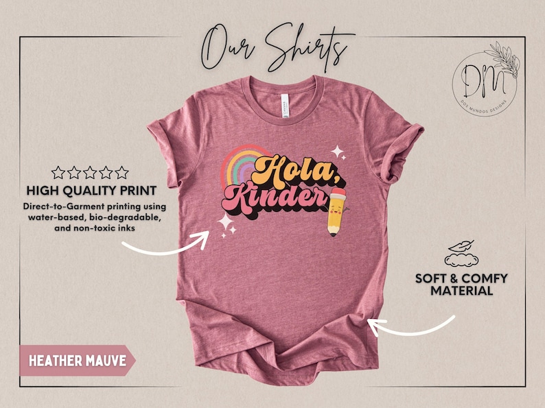 May include: A heather mauve short-sleeve t-shirt featuring a graphic print with the text "Hola, Kinder" in a retro font. The design includes a colorful rainbow, two small four-pointed stars, and a cartoon pencil with a smiling face on the right side.