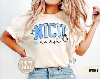May include: An ivory t-shirt featuring the text NICU in large light blue block letters with a black outline. Below, the word nurse is written in black script. Two cartoon penguins, one with a nurse cap and one with a stethoscope, and blue snowflakes decorate the design.