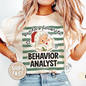 May include: An ivory t-shirt featuring a graphic of Santa Claus with the text "Santa's favorite" above and "BEHAVIOR ANALYST" below. The design is set against a background of horizontal dark green stripes and small star icons.