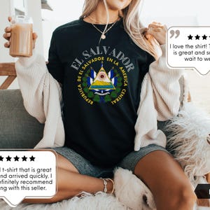 May include: A black short-sleeved t-shirt featuring the coat of arms of El Salvador printed in the center. The design includes the text REPUBLICA DE EL SALVADOR EN LA AMERICA CENTRAL in a circular arrangement around the national emblem.