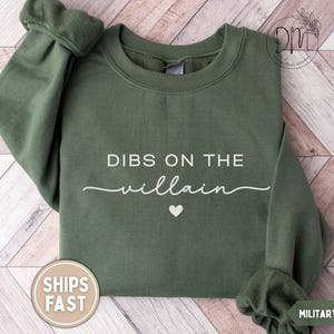 May include: Military green sweatshirt with the text "DIBS ON THE villain" in white script, a small white heart, and the words "SHIPS FAST" on a circular sticker. The sweatshirt has a crew neck and long sleeves.
