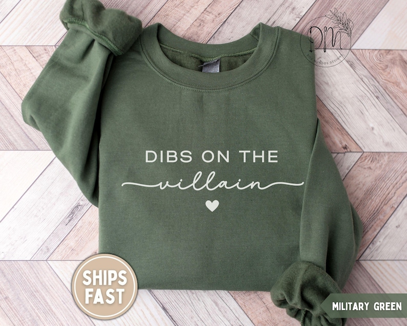 May include: Military green sweatshirt with the text "DIBS ON THE villain" in white script, a small white heart, and the words "SHIPS FAST" on a circular sticker. The sweatshirt has a crew neck and long sleeves.