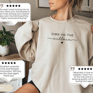 May include: A sand-colored sweatshirt with the text "DIBS ON THE villain" in black script and a small black heart. The sweatshirt is a casual style, suitable for everyday wear.