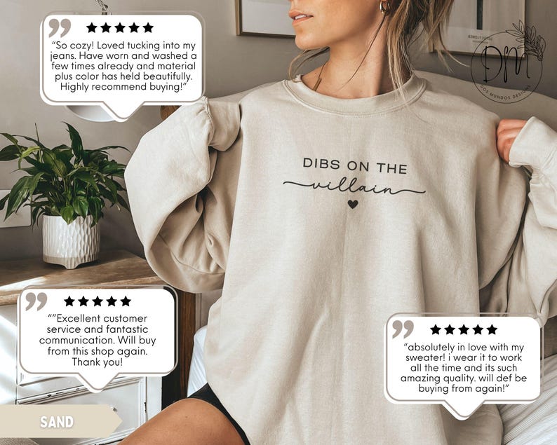 May include: A sand-colored sweatshirt with the text "DIBS ON THE villain" in black script and a small black heart. The sweatshirt is a casual style, suitable for everyday wear.