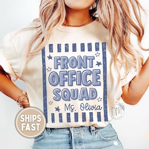 May include: An ivory short-sleeve t-shirt featuring a rectangular blue graphic with vertical stripes and stars. The text reads FRONT OFFICE SQUAD in large blue letters, with the name Ms. Olivia printed in a smaller script font below the text.