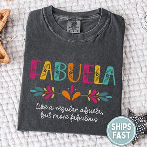 May include: Dark gray t-shirt with the word "ABUELA" in colorful letters. The text "like a regular abuela, but more fabulous" is below. The shirt has a crew neck and is made of a soft material.