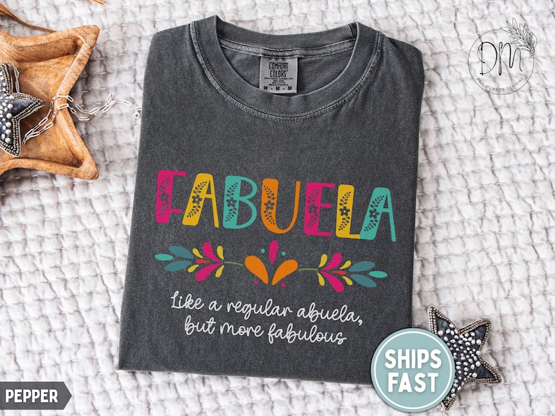 May include: Dark gray t-shirt with the word "ABUELA" in colorful letters. The text "like a regular abuela, but more fabulous" is below. The shirt has a crew neck and is made of a soft material.
