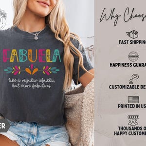 May include: Dark gray t-shirt with the word "ABUELA" in colorful letters. Below the word, it says "Like a regular abuela, but more fabulous." The shirt has a floral design and is printed in the USA.