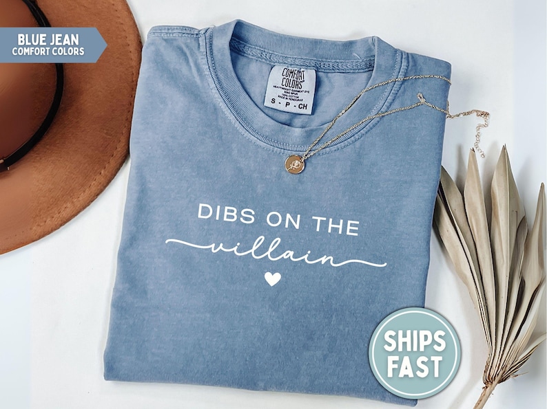 May include: Blue jean Comfort Colors t-shirt with the text "DIBS ON THE villain" in white script and a small heart. A gold necklace and a brown hat are also in the image.