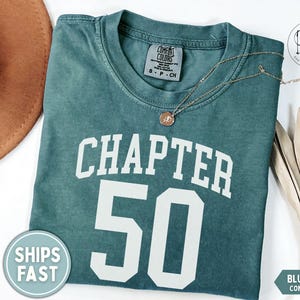 May include: Blue spruce Comfort Colors t-shirt with the text "CHAPTER 50" in white. A gold necklace with a pendant rests on the shirt. A brown hat is visible in the upper left corner.