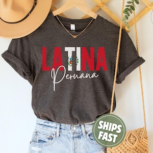 May include: A dark charcoal gray short-sleeve t-shirt featuring the red text LATINA with the Peruvian flag integrated into the letter I, and the word Peruana written in white cursive script below. The shirt has a crew neckline and rolled sleeves.