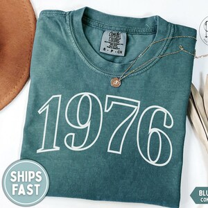 May include: A blue spruce Comfort Colors t-shirt with the number 1976 in white. A gold necklace with a pendant is on the shirt. A brown hat and a palm leaf are in the background. The shirt has a tag that says "Comfort Colors".