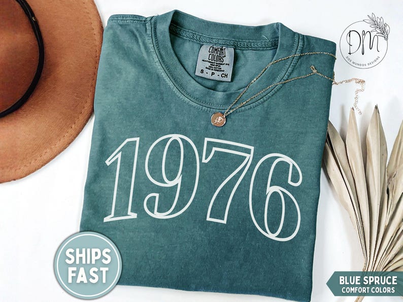 May include: A blue spruce Comfort Colors t-shirt with the number 1976 in white. A gold necklace with a pendant is on the shirt. A brown hat and a palm leaf are in the background. The shirt has a tag that says "Comfort Colors".