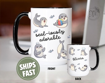 Può includere: Una tazza in ceramica bianca con manico e interno neri, decorata con foche disegnate, piccoli pesci e il testo "seal-iously adorable" sul davanti, e il nome "Olivia" con altre foche e pesci sul retro della tazza.