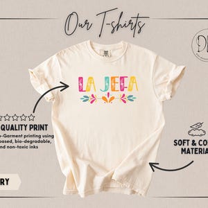 May include: Ivory t-shirt featuring the text "LA JEFA" in vibrant colours and floral designs. The shirt is described as soft and comfy, with high-quality, water-based, and non-toxic print.