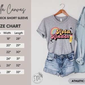 May include: A heather gray crewneck short sleeve t-shirt featuring a colorful graphic with a rainbow, a yellow pencil, and the text "Hola, Kinder" in a retro font. A size chart is provided with measurements in inches for sizes S through 3XL.