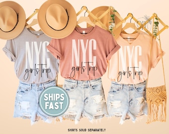 May include: Three t-shirts in gray, coral, and peach, each with "NYC girls trip" printed in white. The shirts are paired with denim shorts and tan hats. A macrame bag hangs on the right. The text "SHIPS FAST" is on a round sticker.