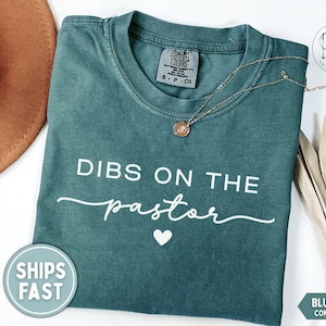 May include: Blue spruce Comfort Colors t-shirt with the text "DIBS ON THE pastor" in white script. A gold necklace with a pendant rests on the shirt. A brown hat and dried palm leaves are also visible.