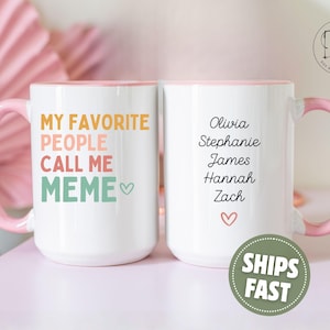 May include: White ceramic mug with a pink handle and rim. The front of the mug reads "MY FAVORITE PEOPLE CALL ME MEME" in colorful text. The back of the mug lists names and a heart symbol.