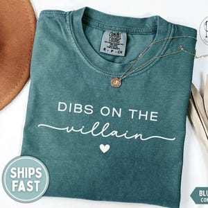May include: Blue spruce Comfort Colors t-shirt with the text "DIBS ON THE villain" in white. A gold necklace with a pendant rests on the shirt. A brown hat and dried plant are in the background.