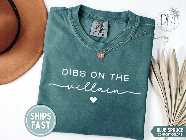 May include: Blue spruce Comfort Colors t-shirt with the text "DIBS ON THE villain" in white. A gold necklace with a pendant rests on the shirt. A brown hat and dried plant are in the background.
