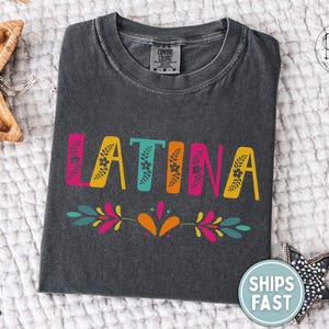 Puede incluir: Camiseta gris oscuro con la palabra "LATINA" en letras coloridas y decoradas con flores. Un dise&ntilde;o floral decorativo est&aacute; debajo de la palabra. La camiseta se muestra sobre una superficie blanca y texturizada.