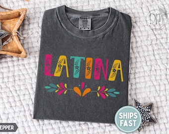 May include: Dark grey t-shirt featuring the word "LATINA" in vibrant, floral-decorated letters. A decorative floral design is below the word. The shirt is displayed on a white, textured surface.