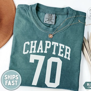 May include: Blue spruce Comfort Colors t-shirt with the text "CHAPTER 70" in white block letters. A gold necklace with a pendant rests on the shirt. A brown hat and a palm frond are in the background.