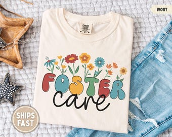 Può includere: T-shirt color avorio con la scritta "FOSTER CARE" in lettere a bolle colorate, con fiori che crescono dalle lettere. La maglietta è abbinata a jeans blu. La maglietta è realizzata in un materiale morbido.