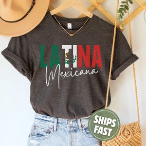 May include: A dark charcoal gray short-sleeve t-shirt featuring the text LATINA in block letters with the Mexican flag colors and emblem integrated into the letter I, and the word Mexicana in white cursive script underneath the main text.