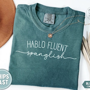 K&ouml;nnte beinhalten: Blaugr&uuml;nes Comfort Colors T-Shirt mit dem wei&szlig;en Text "HABLO FLUENT spanglish". Eine goldene Halskette mit Anh&auml;nger liegt auf dem Shirt. Ein brauner Hut und getrocknete Palmbl&auml;tter sind ebenfalls im Bild.