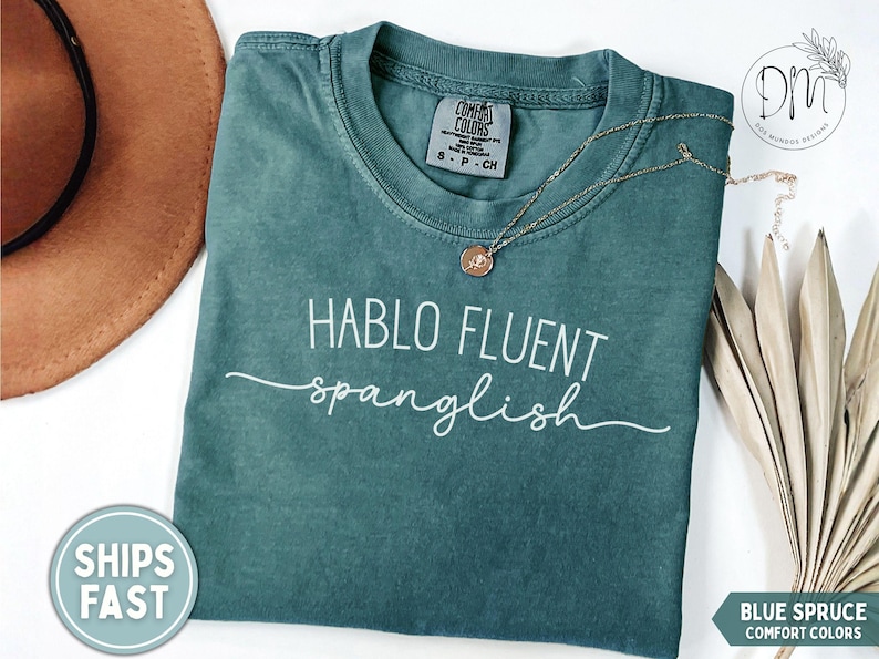 K&ouml;nnte beinhalten: Blaugr&uuml;nes Comfort Colors T-Shirt mit dem wei&szlig;en Text "HABLO FLUENT spanglish". Eine goldene Halskette mit Anh&auml;nger liegt auf dem Shirt. Ein brauner Hut und getrocknete Palmbl&auml;tter sind ebenfalls im Bild.