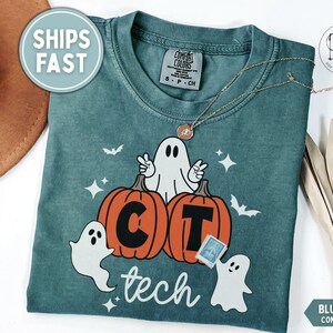 May include: A blue spruce Comfort Colors t-shirt featuring a graphic of three white ghosts and two orange pumpkins. The pumpkins are marked with the letters C and T, and the word tech is printed below in white script. Small bats and stars surround the figures.