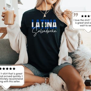 May include: A black short-sleeved t-shirt featuring the text LATINA in bold blue and white letters, with the flag of El Salvador integrated into the design. Below the word LATINA, the word Salvadore&ntilde;a is printed in a white cursive script font.