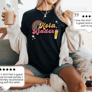 May include: A black t-shirt featuring a colorful graphic with a rainbow, the text "Hola, Kinder" in a retro font, and a small yellow pencil illustration. The design includes four-pointed star accents in white, set against the solid black fabric.