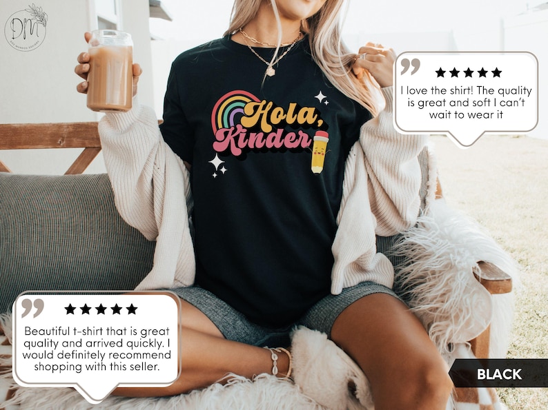 May include: A black t-shirt featuring a colorful graphic with a rainbow, the text "Hola, Kinder" in a retro font, and a small yellow pencil illustration. The design includes four-pointed star accents in white, set against the solid black fabric.