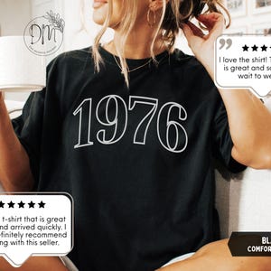 May include: Black t-shirt with the year "1976" in white, outlined font. The shirt is part of the "Comfort Colors" collection. The image also includes customer reviews.