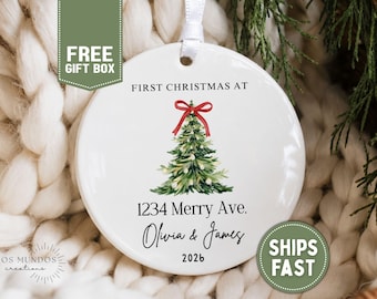 May include: A round white ceramic ornament featuring a watercolor illustration of a green Christmas tree topped with a red bow. The text reads FIRST CHRISTMAS AT, 1234 Merry Ave., Olivia & James, and 2026. A white ribbon is attached to the top.