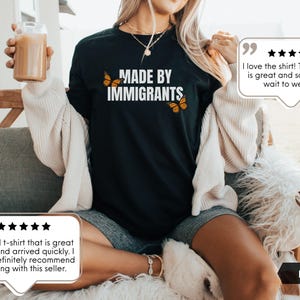 Pu&ograve; includere: Una t-shirt nera a maniche corte con la scritta bianca "MADE BY IMMIGRANTS" sul petto, affiancata da due illustrazioni di farfalle monarca arancioni e nere. Il capo &egrave; abbinato a un cardigan color crema e a pantaloncini grigi.