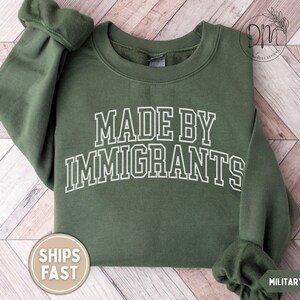 May include: Military green sweatshirt with the text "MADE BY IMMIGRANTS" in white block letters. The sweatshirt has a crew neck and long sleeves. A round sticker with the text "SHIPS FAST" is visible.