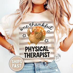 May include: An ivory t-shirt featuring a graphic of a brown turkey with colorful tail feathers centered over horizontal gray stripes. The text reads one thankful PHYSICAL THERAPIST in black script and bold block lettering with small star accents.