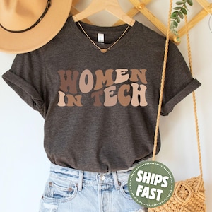 May include: A dark heather gray short-sleeve t-shirt featuring the text "WOMEN IN TECH" in a wavy, multi-toned brown and beige font. The shirt has a crew neckline and is displayed on a hanger with a tan hat and a woven bag visible nearby.