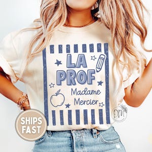 May include: An ivory t-shirt featuring a blue graphic design with vertical stripes, stars, an apple, and a pencil. The text reads LA PROF, Madame Mercier. The design is centered on the chest of the shirt, which is worn with blue denim jeans.