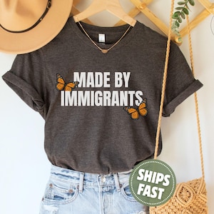 Pu&ograve; includere: Una t-shirt a maniche corte color grigio antracite scuro con la scritta bianca MADE BY IMMIGRANTS stampata al centro. Due farfalle monarca arancioni e nere sono posizionate a sinistra e a destra del testo, aggiungendo un dettaglio colorato.