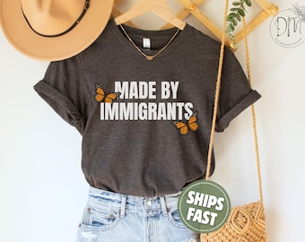 May include: A dark charcoal grey short-sleeve t-shirt featuring the white text MADE BY IMMIGRANTS printed in the centre. Two orange and black monarch butterflies are positioned on the left and right sides of the text, adding a colourful detail.